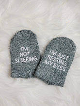 Load image into Gallery viewer, One pair of green socks are laying on a white fuzzy rug. The socks are folded so that you can see the soles, where there is text in white. On one sock it says I'm not sleeping. On the other it says I'm just resting my eyes.
