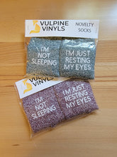 Load image into Gallery viewer, Two packages of socks sit on a wooden table to show how they are packaged. They are folded in a clear plastic bag with a paper topper. The topper says Vulpine Vinyls Novelty Socks. The socks are folded so that you can read the sayings on them.