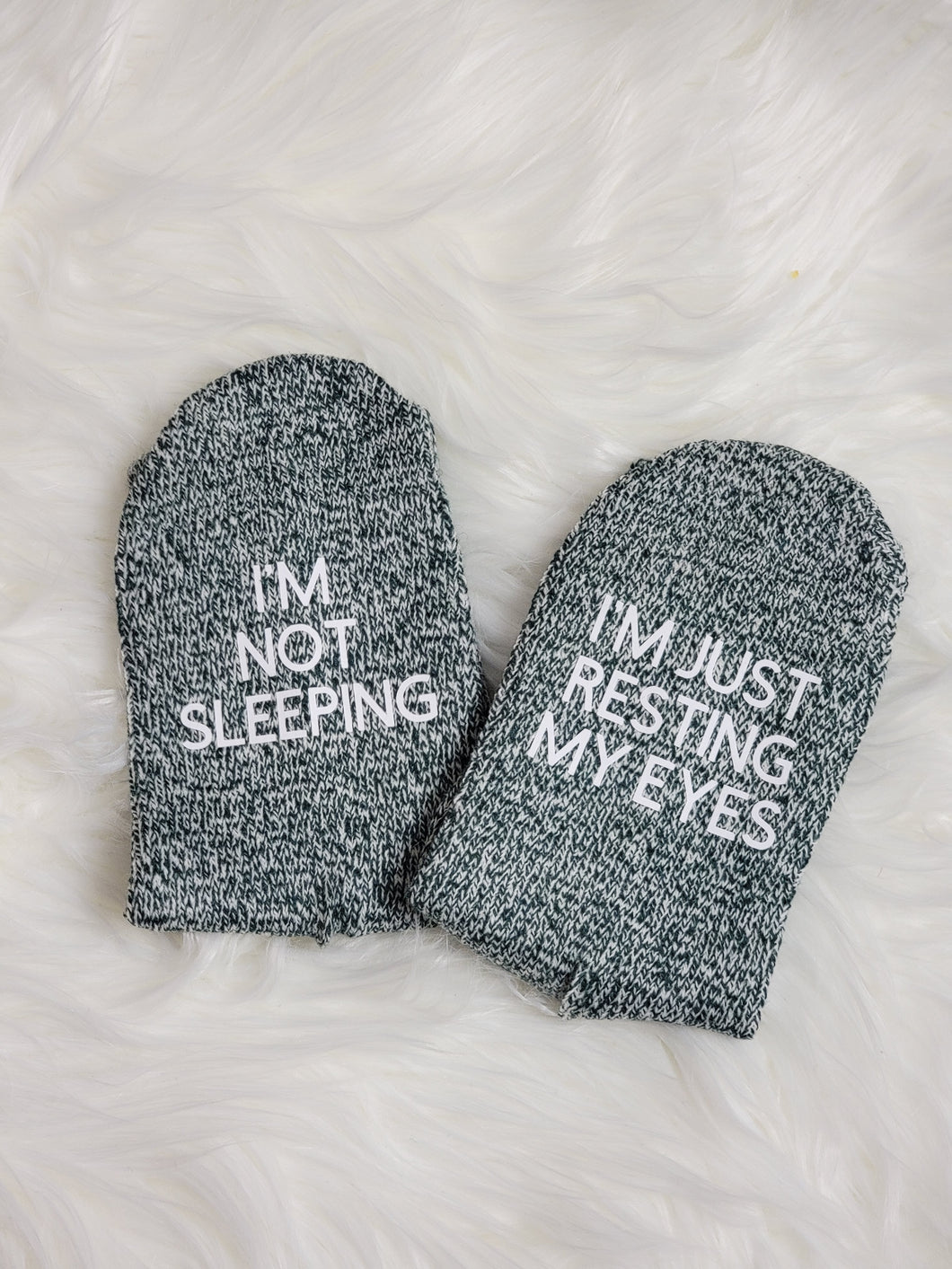 One pair of green socks are laying on a white fuzzy rug. The socks are folded so that you can see the soles, where there is text in white. On one sock it says I'm not sleeping. On the other it says I'm just resting my eyes.