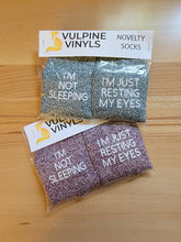 Load image into Gallery viewer, Two packages of socks sit on a wooden table to show how they are packaged. They are folded in a clear plastic bag with a paper topper. The topper says Vulpine Vinyls Novelty Socks. The socks are folded so that you can read the sayings on them.

