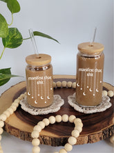 Load image into Gallery viewer, Tw0 cup sit on tan coasters on a wooden lazy Susan. The cups have bamboo lids and glass straight straws. The glasses are filled with coffee, and have the text manifest that shit with stars and moons dangling down from the text in white.
