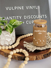 Load image into Gallery viewer, A glass can shaped cup sits on a tan coaster on a wooden lazy Susan. The cup has a bamboo lid and a glass straight straw. The glass is filled with coffee, and has the text manifest that shit with stars and moons dangling down from the text.
