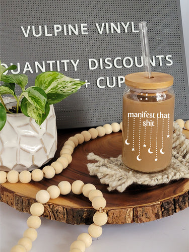 A glass can shaped cup sits on a tan coaster on a wooden lazy Susan. The cup has a bamboo lid and a glass straight straw. The glass is filled with coffee, and has the text manifest that shit with stars and moons dangling down from the text.