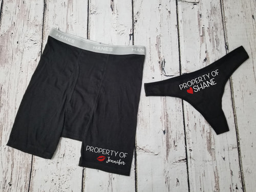 Men's Black boxer briefs and a black thong. The design on the men's boxer briefs says Property of Jennifer with red kiss lips next to the name. The women's thong says property of Shane with a red heart next to the name.