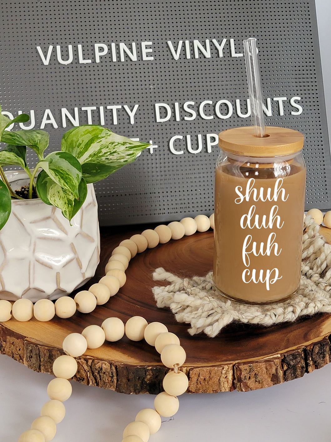 A glass can shaped cup sits on a blue coaster on a wooden lazy Susan. The cup has a bamboo lid and a glass straight straw. The glass is filled with coffee, and has the text Shuh Duh Fuh Cup on it in a white cursive font.